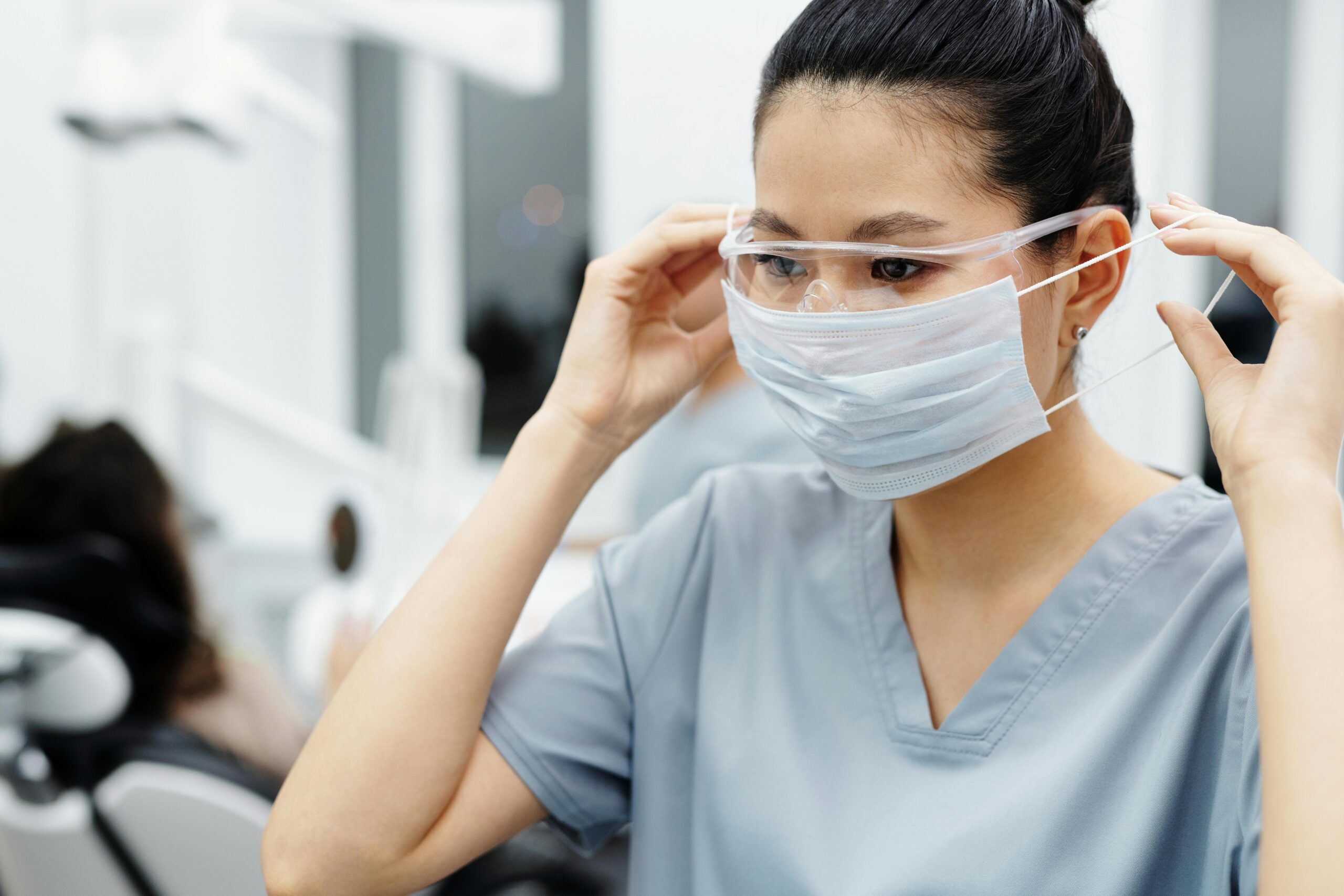 Dental nurse in scrubs adjusts goggles and face mask in a clinic.